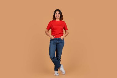 Genç bir ortadoğulu kadın bronz bir arka planın önünde kırmızı tişört ve mavi kot pantolon giyerek duruyor. Gülümsüyor ve elleri cebinde. Beyaz spor ayakkabı giyiyor..