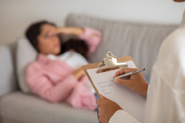 Bir terapist elinde bir not panosu ve kalemle oturur ve hasta arka planda bir kanepede yatarken not alır. Hasta sıkıntılı ve odakta değil gibi görünüyor..