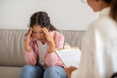 Bu görüntüde bir kadın kanepeye oturmuş, başını ellerinin arasına almış, sıkıntılı görünüyor. Terapi seansında ve bir terapist karşısında elinde bir not panosuyla oturuyor.