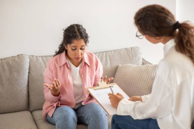 Pembe gömlekli bir kadın kanepede oturuyor ve yanında oturan bir terapistle konuşuyor. Kadın bir şeyleri açıklarken terapist bir not defterine not alıyor.