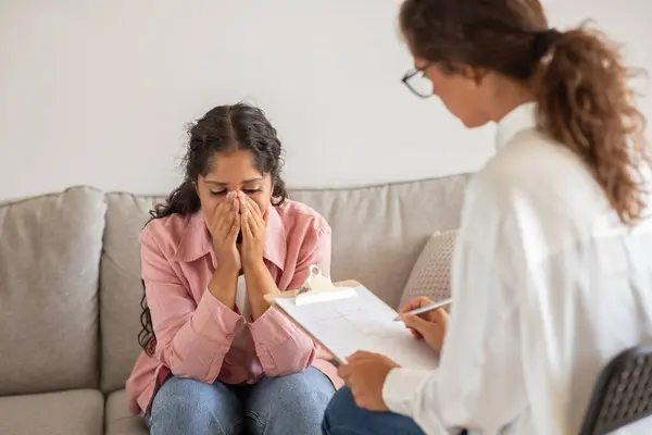 stock image A woman is sitting on a couch in an office, crying while being counseled by a therapist. The therapist is sitting to her right, holding a clipboard with a pen in her hand, seemingly taking notes.