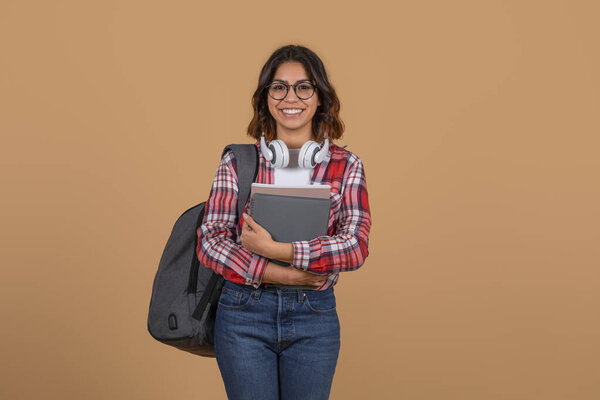 Молодая студентка с Ближнего Востока в красно-белой клетчатой рубашке, с пачкой книг и рюкзаком. Она в очках и улыбается в камеру. На заднем плане простой цвет загара.