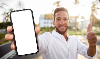 Bir adam dışarıda duruyor, elinde boş bir ekranla akıllı bir telefon tutuyor ve onayı veriyor. Gülümsüyor ve doğrudan kameraya bakıyor..