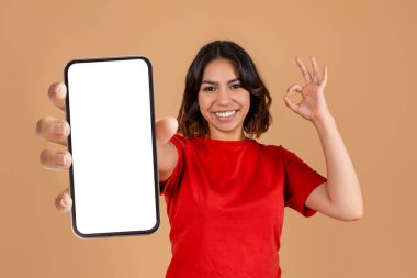 Genç bir ortadoğulu kadın gülümsüyor ve önünde boş bir ekranla akıllı bir telefon tutuyor. Kırmızı bir gömlek giyiyor ve diğer eliyle 