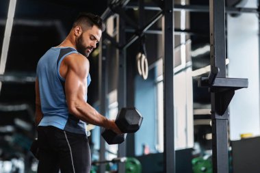 Spor salonunda bir adam egzersiz sırasında dambılı kaldırır. Kolsuz bir gömlek ve siyah pantolon giyiyor..