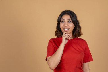 Kahverengi saçlı, orta doğulu bir kadın kırmızı bir tişört giyiyor ve yukarı ve sola bakıyor. Düşünceli bir ifadesi var ve elini çenesine dayayıp uzayı kopyalıyor.