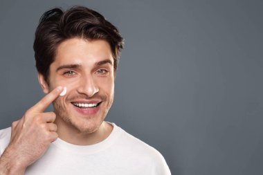 Sabah rutini. Yanak, gri arka plan üzerinde yüz kremi uygulayan genç adam
