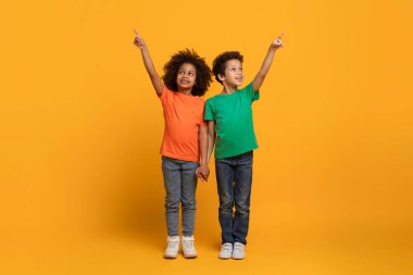 Afro-Amerikalı iki çocuk, turuncu tişörtlü bir kız ve yeşil gömlekli bir oğlan, parlak sarı arka planda duruyor. İki çocuk da sağ işaret parmaklarıyla yukarı bakıyor.