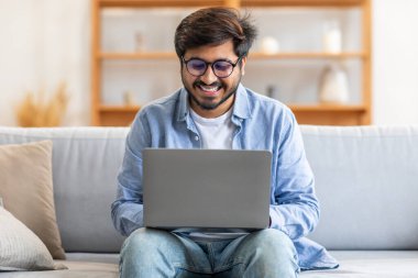 Kapalı bir koltukta oturan Hintli bir adam, dizüstü bilgisayarını iş için ya da boş zaman aktiviteleri için kullanmaya dalmış. Ekranla etkileşime girerken odaklanmış ve konsantre olmuş görünüyor..