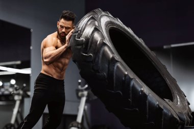 Tişörtsüz ve kaslı bir adam spor salonunda ağır bir lastiği kaldırıyor..