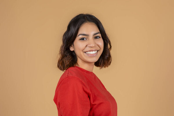 Молодая средневосточная женщина с темными каштановыми волосами, улыбающаяся и смотрящая прямо в камеру. Она одета в ярко-красную рубашку с длинными рукавами и стоит перед сплошным бежевым фоном.