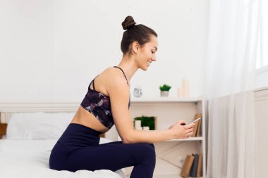 Aktif giyimli bir kadın yatağın kenarında oturuyor ve telefonuna bakıyor..