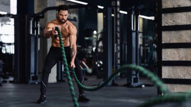 Kaslı bir adam spor salonunda savaş halatlarıyla çalışıyor. O, siyah şort ve ayakkabı giyiyor. Antrenmanına odaklanmış durumda..