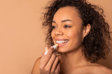 Kıvırcık kahverengi saçlı genç siyah bir kadın gülümsüyor ve dudaklarına dudak kremi sürüyor. O doğal bir makyaj görünümü giyiyor ve açık bir teni var. Arka plan yumuşak bir bej rengi..