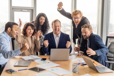 Çeşitli iş adamları modern bir ofisteki bir konferans masasının etrafında toplanarak başarıyı kutluyorlar. Elleri havada, gülümsüyor ve tezahürat yapıyorlar..