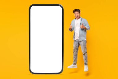 Genç bir adam boş beyaz bir ekranla büyük bir akıllı telefonun önünde havaya zıplıyor, iki eliyle baş parmağını kaldırıyor ve yüzünde kocaman bir gülümseme var. Arka plan parlak sarı.