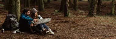 Genç bir çift orman zemininde, yüksek ağaçlar ve düşen yapraklarla çevrili olarak oturuyor. Bir harita üzerinde çalışıyorlar, muhtemelen bir sonraki maceralarını planlıyorlar, kopyalama alanı.