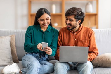 Gülümseyen bir Hintli çift modern oturma odalarında rahat bir koltuğa oturmuş, birlikte vakit geçiriyorlar. Kadın akıllı telefonuna bakarken, adam dizüstü bilgisayar kullanıyor.