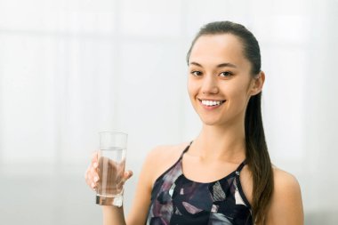 Spor kıyafetleri içinde bir kadın bir bardak su tutar, antrenmandan sonra mutlu bir şekilde gülümser..