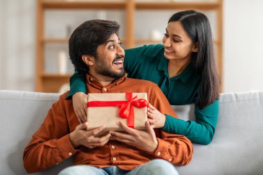 Hintli bir adam bir kanepede oturuyor, yanında bir kadın otururken bir hediye kutusu tutuyor. Kadın ve erkek içeride, arka planda tarafsız bir dekor var..