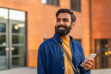 Hintli sakallı adam elinde akıllı bir telefonla büyük bir tuğla binanın önünde duruyor ve gülümsüyor. Sıradan mavi bir gömlek giymişti ve mutlu ve mutlu görünüyordu.