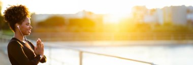 Yoga açık havada. Sabahları dışarıda meditasyon yapan gülümseyen siyahi kadın portresi, kapalı gözlerle Namaste jesti yapan ve kulaklıkla müzik dinleyen,