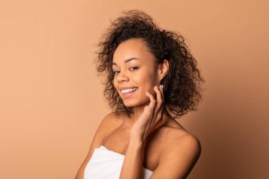 Kıvırcık kahverengi saçlı Afro-Amerikan bir kadının portresi. O gülümsüyor ve beyaz bir havlu giyiyor. Arka plan açık kahverengi bir renktir. Fotoğrafın sıcak ve davetkar bir havası var..