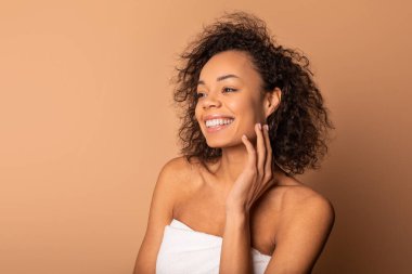 Kıvırcık kahverengi saçlı, gülümseyen Afro-Amerikan kadın beyaz bir havlu giyiyor ve yana bakıyor. O, eliyle yanağına dokunuyor. O, düz bir kahverengi arka planda duruyor..