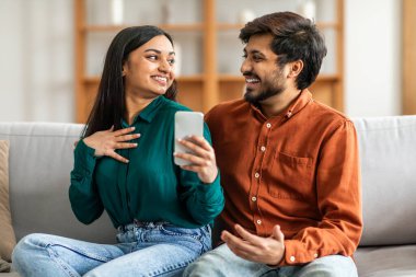 Hintli bir adam ve kadın bir koltukta oturuyorlar, ikisi de ellerinde tuttukları bir cep telefonuyla. Ekrana odaklanmış görünüyorlar, belki bir video izliyorlar ya da sosyal medyada birlikte geziniyorlar..