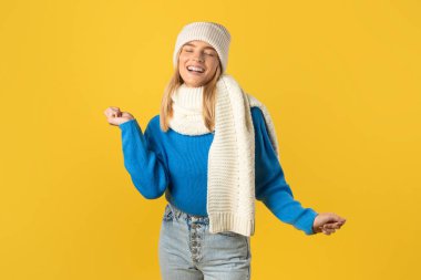 Mavi kazaklı, beyaz örgü şapkalı ve eşarplı neşeli genç bir kadın parlak sarı bir arka planda neşeyle dans ediyor. Gözleri kapalı ve bu anın tadını çıkarıyor gibi görünüyor..