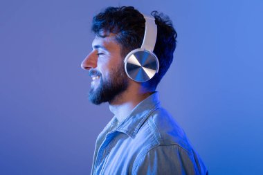 Sakallı bir adam kulaklık takıyor, müzik dinlemeye dalmış. Yüzünde ciddi bir ifadeyle sese odaklanmış görünüyor..