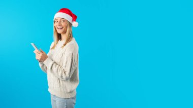 Fotoğrafta, elinde cep telefonu olan ve Noel Baba şapkası takan bir kadın görülüyor. Telefonu kullanıyor gibi görünüyor, muhtemelen selfie çekiyor ya da birisine mesaj atıyor, boş bir alan.