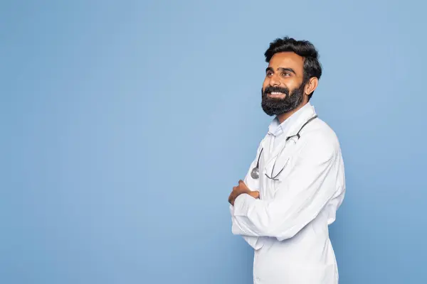 Stock image A bearded Indian doctor wearing a white coat and stethoscope smiles while standing confidently against a blue studio background. His arms are crossed, conveying professionalism and warmth.