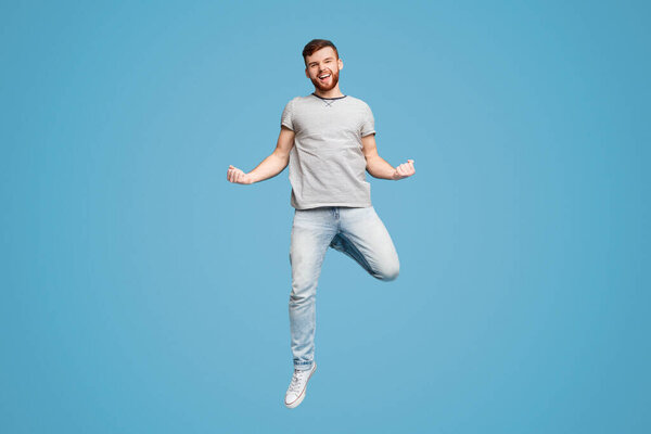 Празднование успеха. Возбужденный молодой рыжеволосый мужчина тысячелетия прыгает и жестикулирует на фоне голубой студии