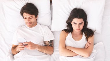 Uzun siyah saçlı genç bir kadın kollarını kavuşturmuş, üzgün görünüyor. Erkek partneri onun yanında uzanmış, telefonunu kullanıyor. Sahne potansiyel bir ilişki sorununu gösteriyor.