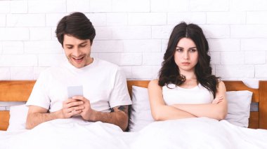 Bir adam yatakta uzanıyor, gülümsüyor ve telefonuna bakıyor. O, beyaz bir tişört giyiyor. Yanında bir kadın yatıyor, kolları çapraz ve başka yöne bakıyor. Mutsuz görünüyor..