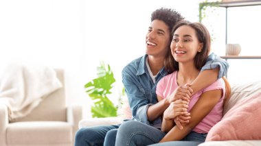 Afro-Amerikalı genç çift oturma odasında bir kanepede oturuyor. Bakıyorlar ve gülümsüyorlar, rahatlamış ve mutlu görünüyorlar, kopyalama alanı.