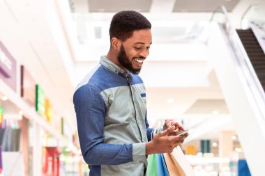 Genç bir Afro-Amerikan adam bir alışveriş merkezinde duruyor, bir elinde alışveriş torbaları diğer elinde telefonuna bakıyor. Cihazında bir şeyi tarıyor veya kontrol ediyor gibi görünüyor.