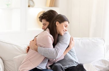 Bir erkek ve bir kadın beyaz bir kanepede oturuyor, kucaklaşarak kucaklaşıyorlar. İlişkilerinde, sevgi ve şefkat ifadeleriyle rahat ve samimi görünürler..