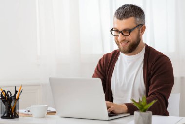 Masada oturan bir adam dizüstü bilgisayarının ekranına odaklanmış. Ekrana dikkatle bakarken elleri klavyeye yazı yazıyor. Çalışma alanı düzenlidir.