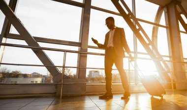 Zenci bir iş gezgini havaalanında yanında bavuluyla telefonuyla konuşuyor. Ortam hareketli ve profesyonel, yoğun bir seyahat günü, güneş parlaması