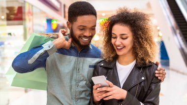 Afro-Amerikalı kadın ve erkek, mağazalar ve diğer müşterilerle çevrili hareketli bir alışveriş merkezinde dolaşırken telefonlarıyla meşgul oluyorlar, cihazlarına odaklanıyorlar, muhtemelen fiyatlarını kontrol ediyorlar.