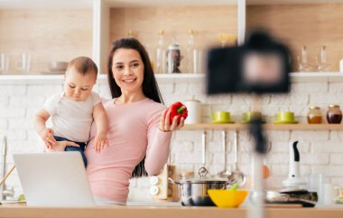 Bir anne, bebeğini kucağında tutarken ve video blogu çekerken modern bir mutfakta yemek yaparken görülüyor. Kameraya dolmalık biber gösteriyor. Tarifini ya da yemek tavsiyelerini paylaştığını gösteriyor..