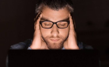 Gözlük takan bir adam gece geç saatlerde dizüstü bilgisayarı üzerinde çalışırken yoğun bir şekilde konsantre olur. Elleri başının üstünde, derin düşüncelere veya strese işaret ediyor..