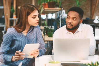 İki meslektaş bir dizüstü bilgisayara bakarken iş hakkında konuşur. Kadının elinde bir not defteri var. Bitkilerin ve doğal ışığın olduğu bir ofisteler..