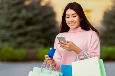 Asyalı bir kadın bir elinde alışveriş çantası ve kredi kartı tutarken diğer elinde cep telefonu kullanırken görülüyor. Birden fazla iş yapıyor gibi görünüyor, muhtemelen mesajlarını kontrol ediyor.