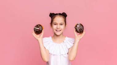 Çikolata kaplı çörek tutan ve pembe stüdyo arka planda gülümseyen mutlu küçük kız