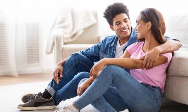 Oturma odasında yerde oturan Afro-Amerikalı genç çift. İkisi de gülümsüyor ve birbirlerine bakıyorlar. Arka planda bir kanepe var ve oda modern bir şekilde dekore edilmiş..