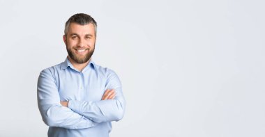 Mavi gömlekli bir adam rahatça poz veriyor, rahat bir şekilde kameraya doğru gülümsüyor. Anın tadını çıkarıyor gibi görünüyor ve sıradan bir poz veriyor, panorama