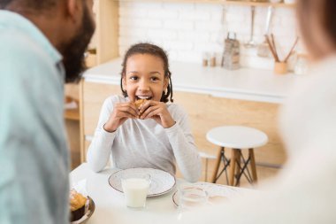 Genç Afrikalı Amerikalı bir kız mutfaktaki masada oturup aile anlarını ve babalığını sergilerken mutlu bir şekilde sandviç yiyor.